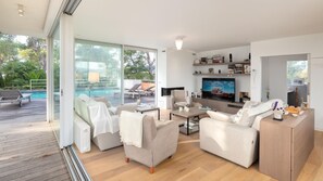 Ein modernes Wohnzimmer mit großem Fernseher, bequemen Sitzmöglichkeiten, Blick auf einen Pool und einer Holzterrasse.