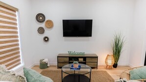 Una sala de estar moderna con un televisor de pantalla plana, un letrero que dice 'Welcome', una mesa de centro redonda y una silla de mimbre.