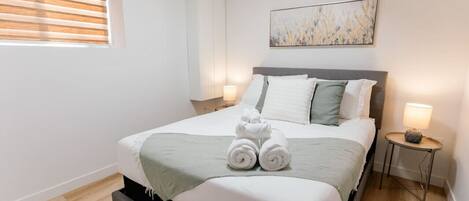 A neatly made bed with a grey headboard, a bedside table with a lamp, and a window with blinds.