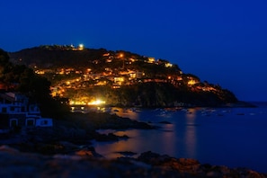 Un pueblo costero de noche con edificios iluminados en una colina que domina el mar.