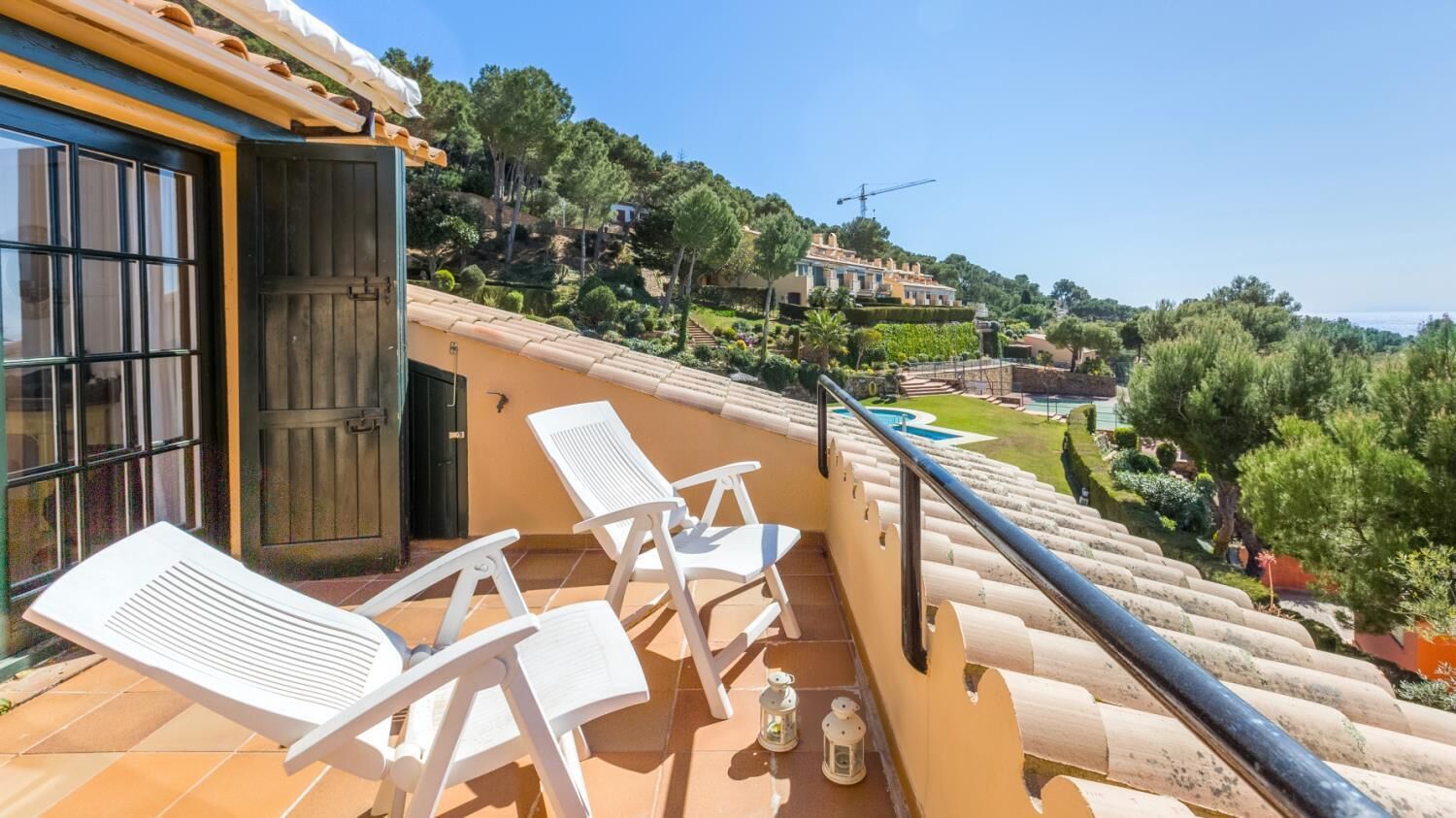 A balcony with white chairs, a railing, and a view of a landscaped garden and buildings in the distance.