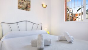 Una cama bien hecha con sábanas blancas y dos toallas enrolladas, un cabecero de metal, una lámpara fijada a la pared y una ventana con vistas al exterior.