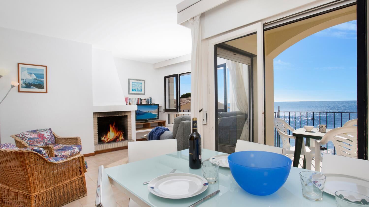 Een balkon met een eettafel, stoelen en uitzicht op zee. In de woonkamer is een open haard en een bank.