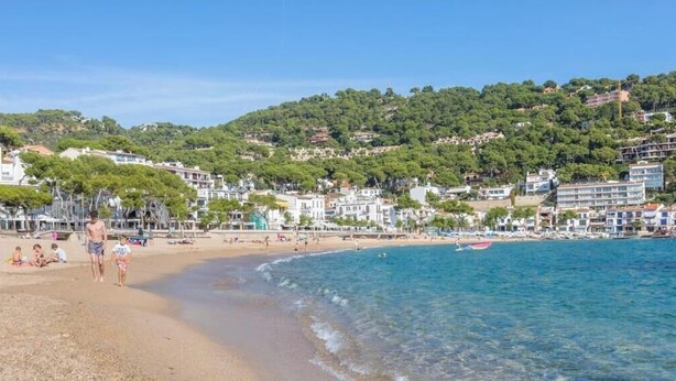 Una playa con agua cristalina, una orilla arenosa y edificios al fondo.