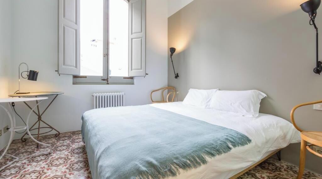 Un dormitorio con cama, un radiador, una ventana con contraventanas abiertas, un escritorio con lámpara y una silla.