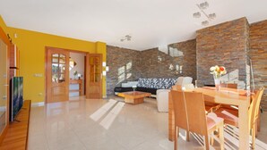 Un salon moderne avec un mur jaune, un mur d’accent en briques, un canapé, une table à manger avec des chaises et un téléviseur.