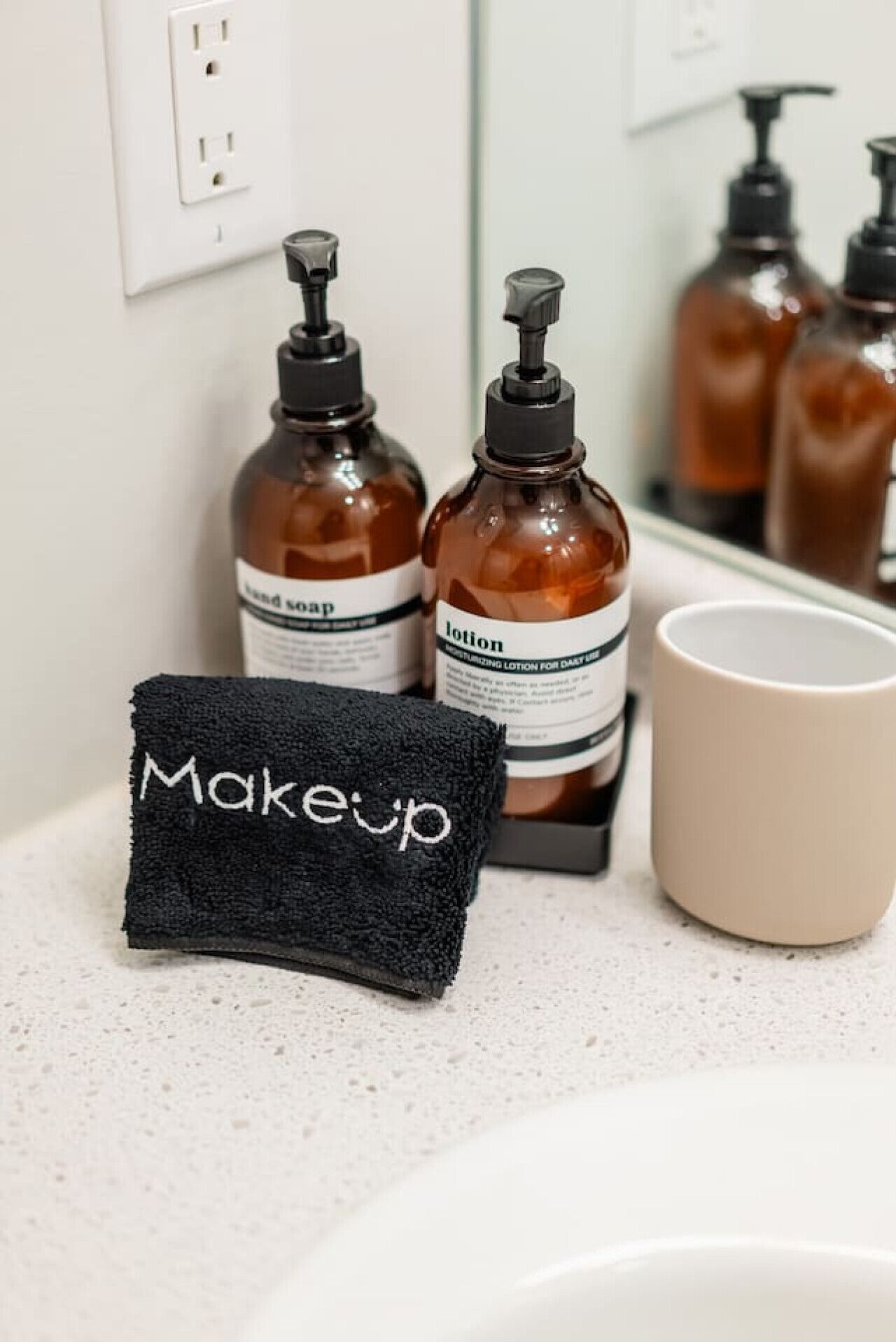 Una encimera de baño con dos botellas de loción, una toalla negra con la etiqueta 'Maquillaje' y una taza color beige.