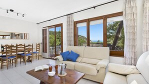 A spacious living area with a sofa, coffee table, and chairs. Large windows offer a view of greenery and a building. - Carena 1 in Llafranc (Palafrugell)
