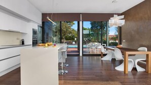 Una cocina moderna con una isla central, un comedor con sillas blancas y vistas a una piscina exterior.
