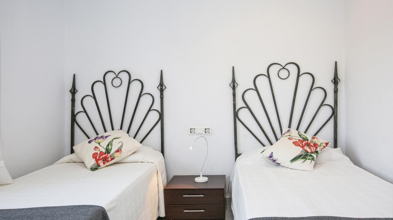 Een slaapkamer met twee bedden, elk met een bloemenkussen en een nachtkastje met een lamp.