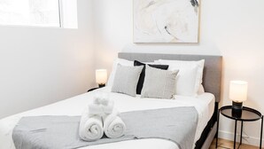 Ein ordentlich bezogenes Bett mit gestreiften Kissen, einer grauen Decke und aufgerollten Handtüchern. Darüber an der Wand hängt ein Gemälde, und auf jeder Seite des Bettes stehen zwei Nachttischlampen.
