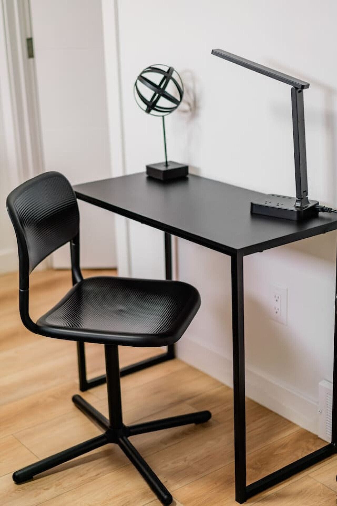 Un bureau noir avec une lampe au design moderne, une chaise noire à dossier en maille et une sphère décorative accrochée au mur.