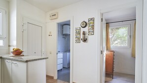 A kitchen with white cabinets, a countertop with a fruit bowl, a window with curtains, and a door leading to a bathroom. - Cliper C 3 in Llafranc (Palafrugell)