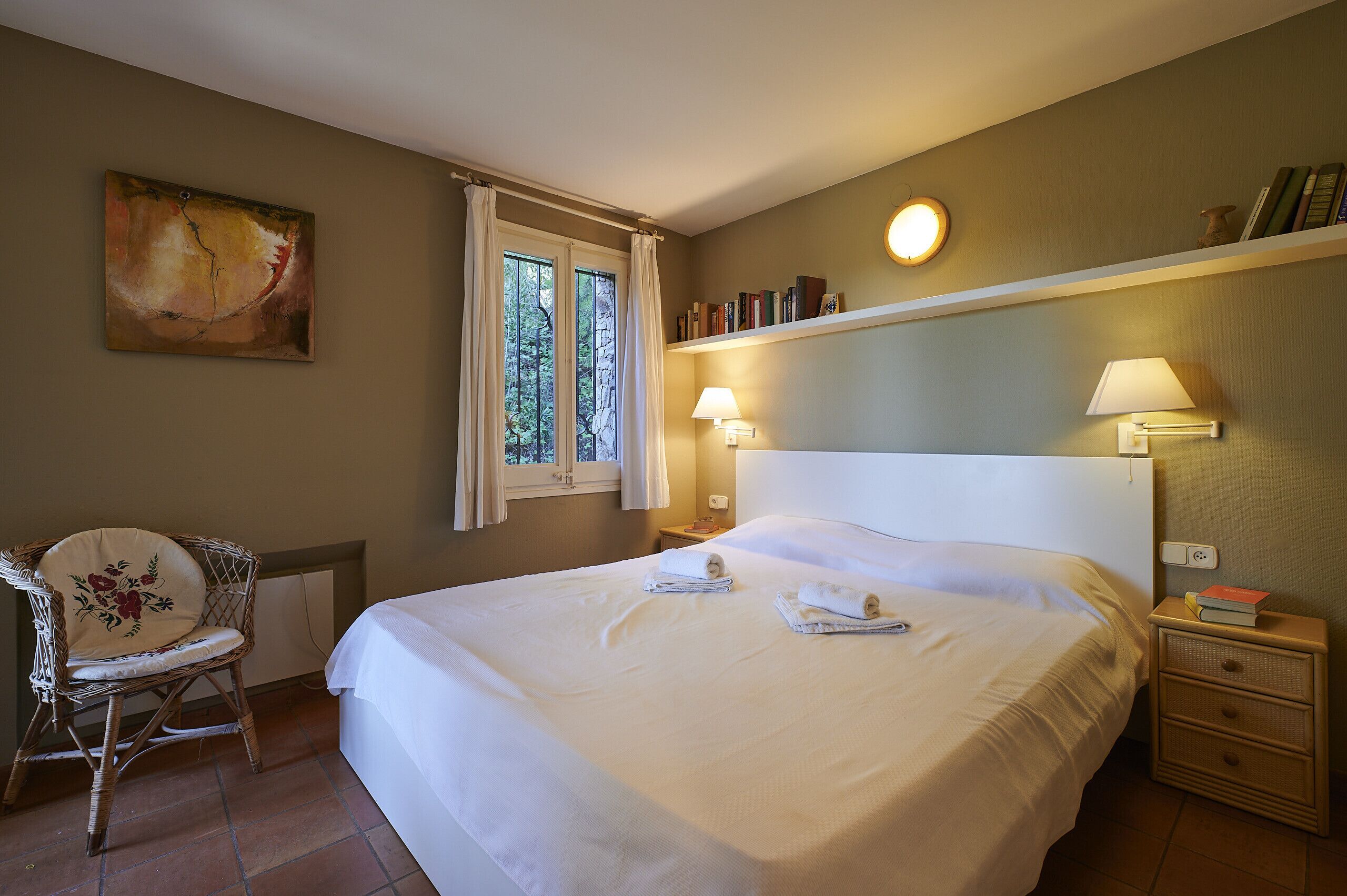 A neatly made bed with two towels, a wicker chair, a bedside table with a lamp, and a bookshelf with books.