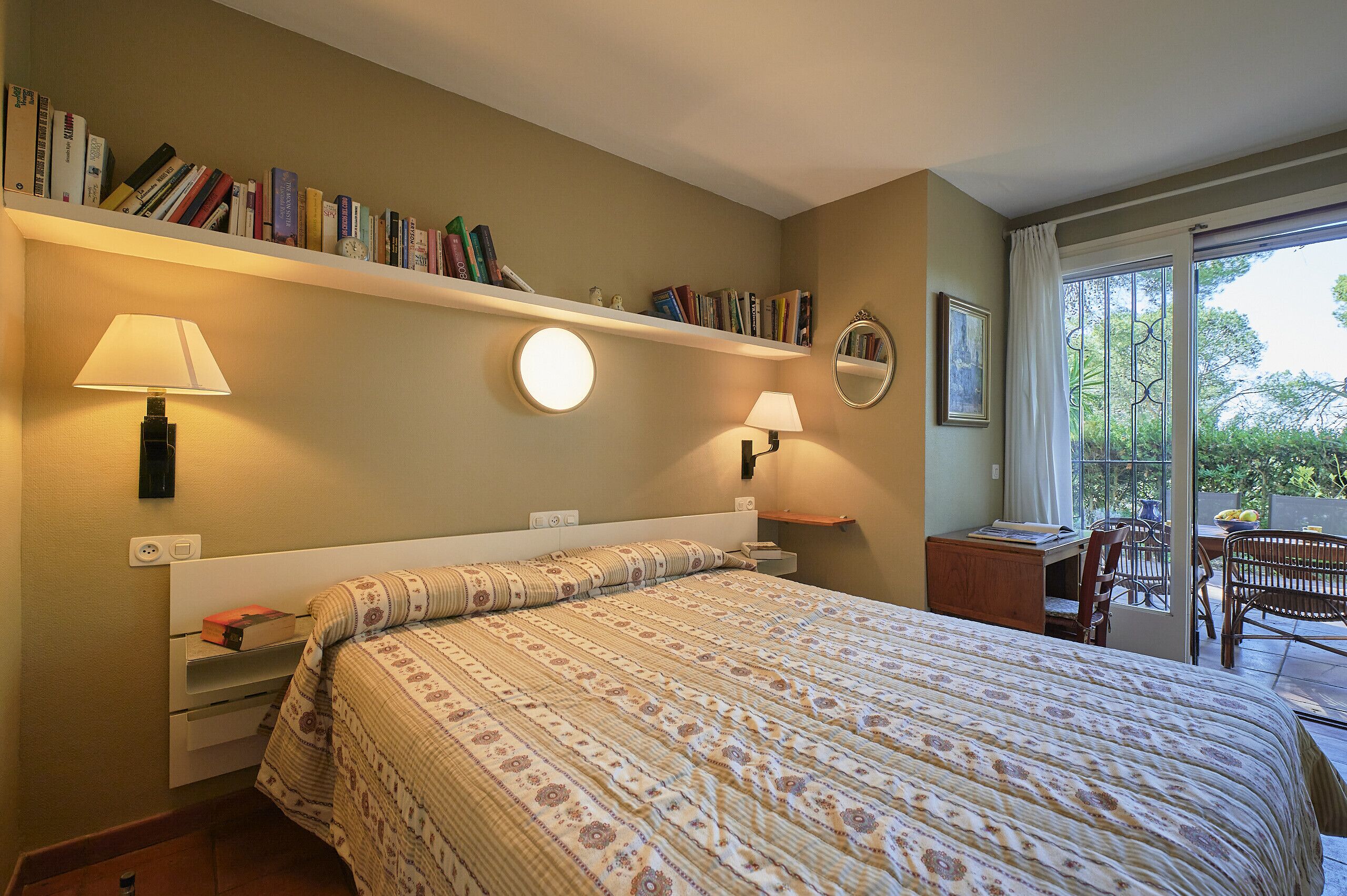 A bedroom with a large bed, two bedside lamps, a bookshelf, a desk, and a view of the outdoors through a sliding glass door.