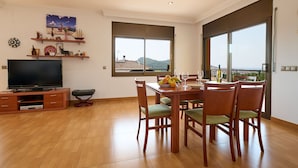 Un vaste espace de vie comprenant un téléviseur à écran plat, un meuble de rangement en bois, une table à manger avec des chaises et de grandes fenêtres offrant une vue sur la mer et les collines.