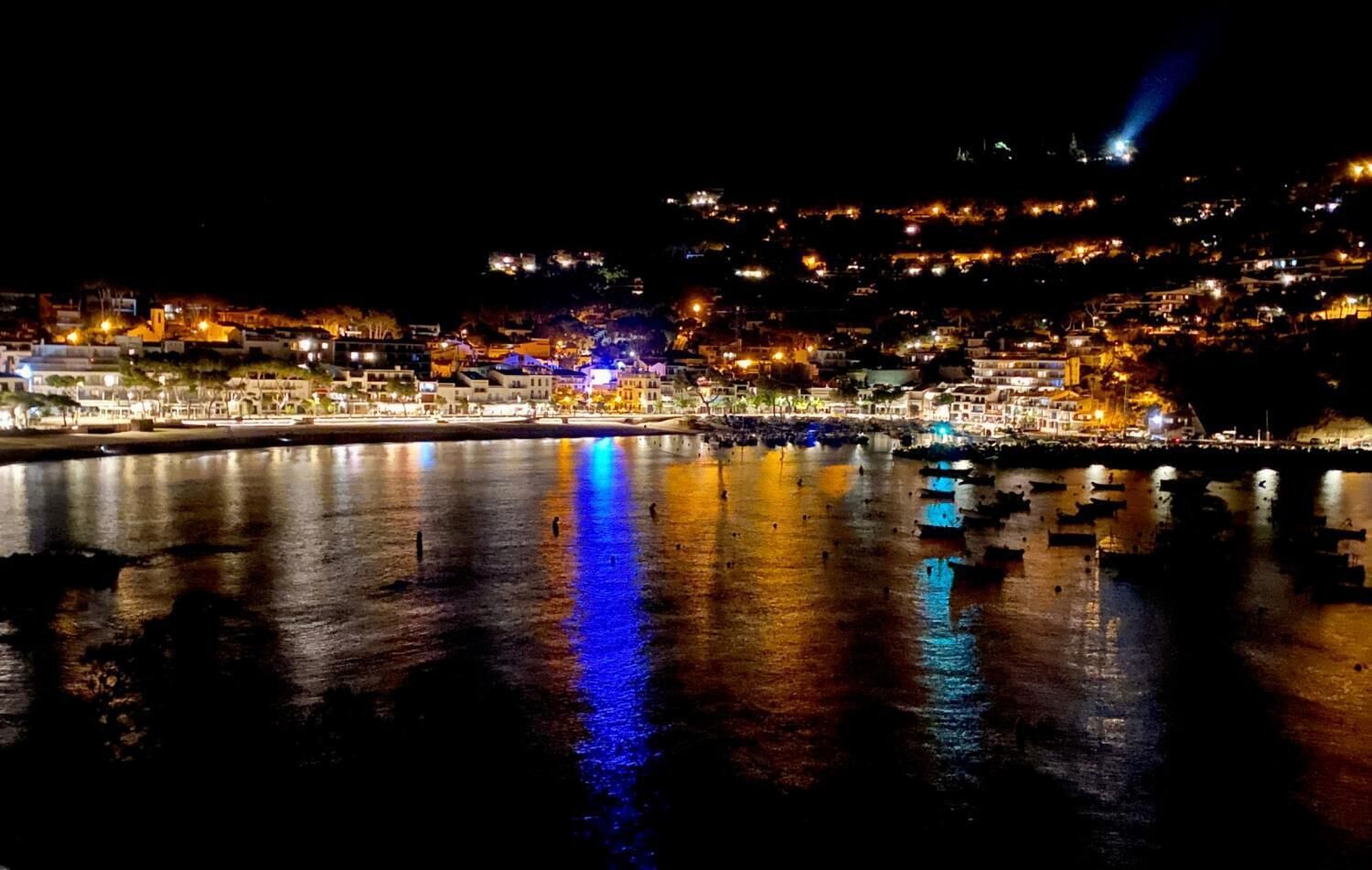 A nighttime coastal cityscape with illuminated buildings and boats on calm waters.