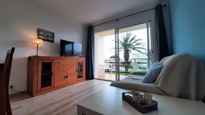 Un soggiorno moderno con un divano, un mobile in legno, una televisione e un balcone che offre vista su palme e un edificio.