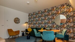 Un salon moderne avec une table ronde en bois, des fauteuils de couleur turquoise et jaune moutarde, un canapé vert et un papier peint à motifs floraux.