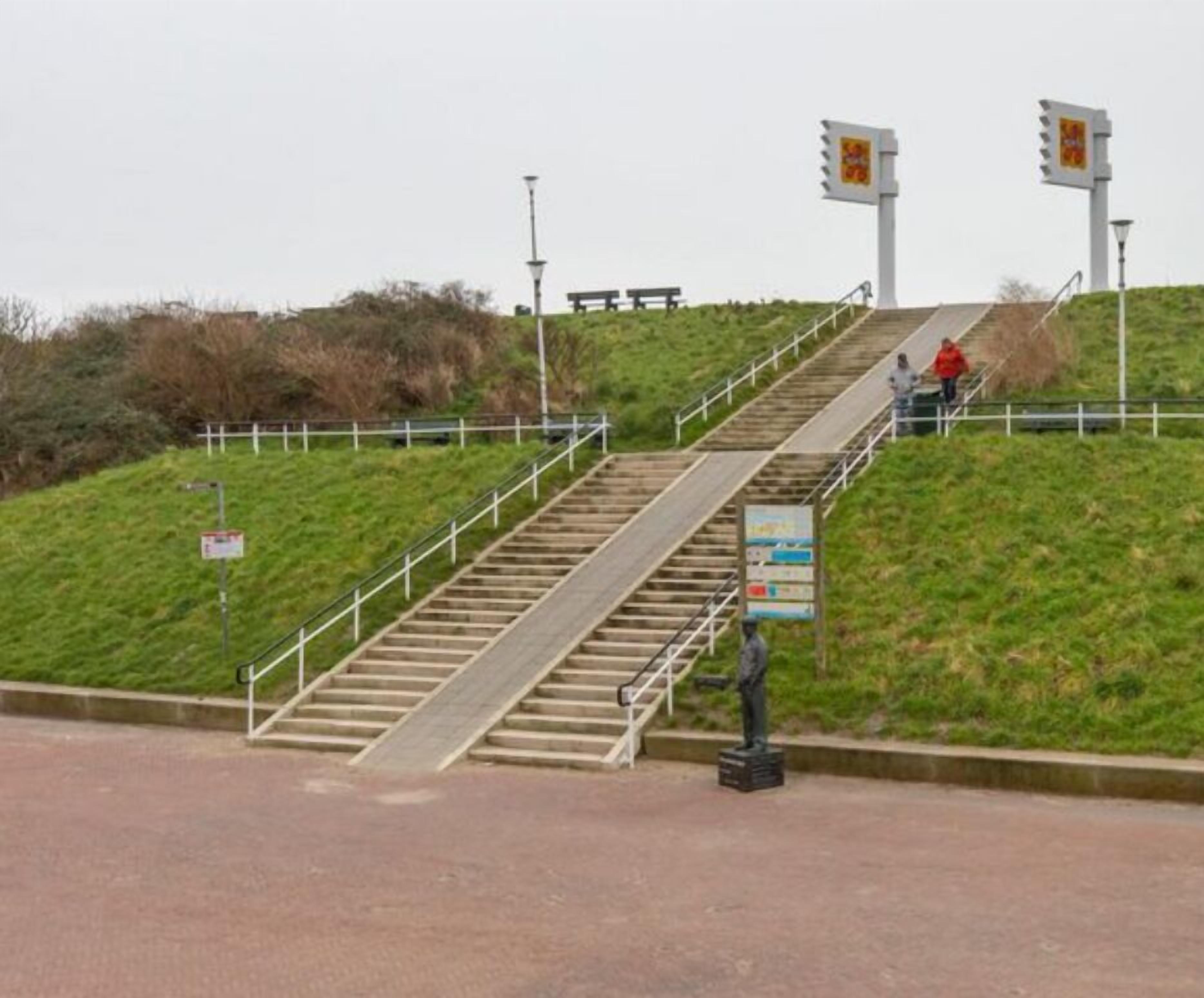 A set of outdoor stairs leading up a grassy hill with a metal railing, a statue, and informational signs.