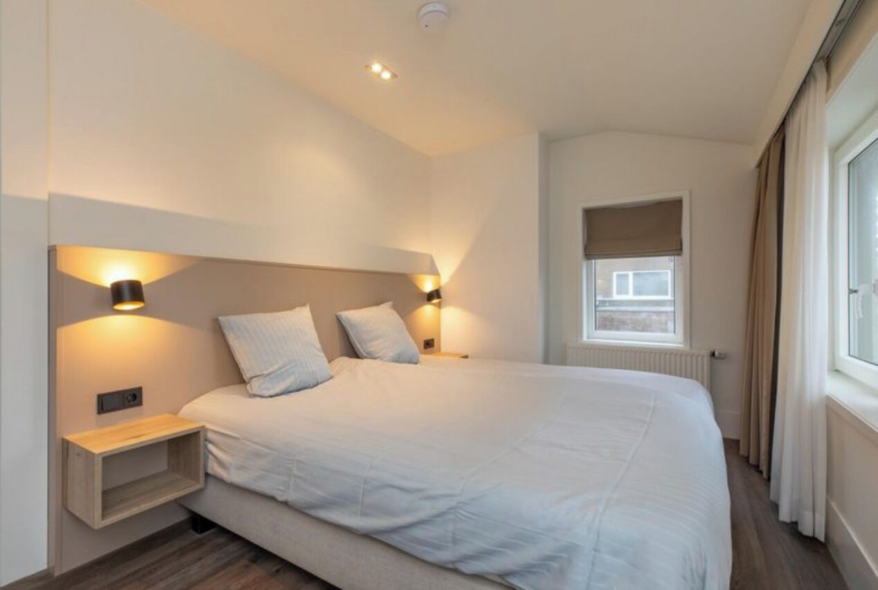 A modern bedroom with a large bed, bedside lamps, a small wooden nightstand, and a window with curtains.