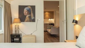 Habitación de hotel con cama, mesita de noche, lámpara, un cuadro y vista al área de comedor.