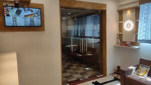 A modern living room with a television mounted on the wall displaying a video, a glass door leading to a dining area, a sofa, and a decorative wall piece. - HOTEL SAVERA (Ahmedabad)
