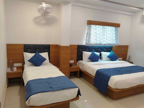 Habitación de hotel con dos camas, cada una con cabecero de madera, ropa de cama blanca y detalles azules. Se ve una ventana con cortinas translúcidas, una mesita de noche con una botella y un ventilador montado en la pared.