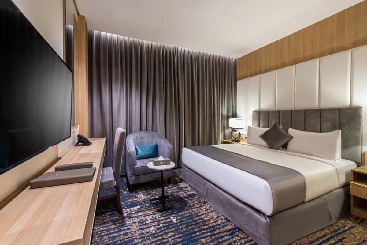 A hotel room with a large bed, a desk with a flat-screen TV, a chair, and a small table.
