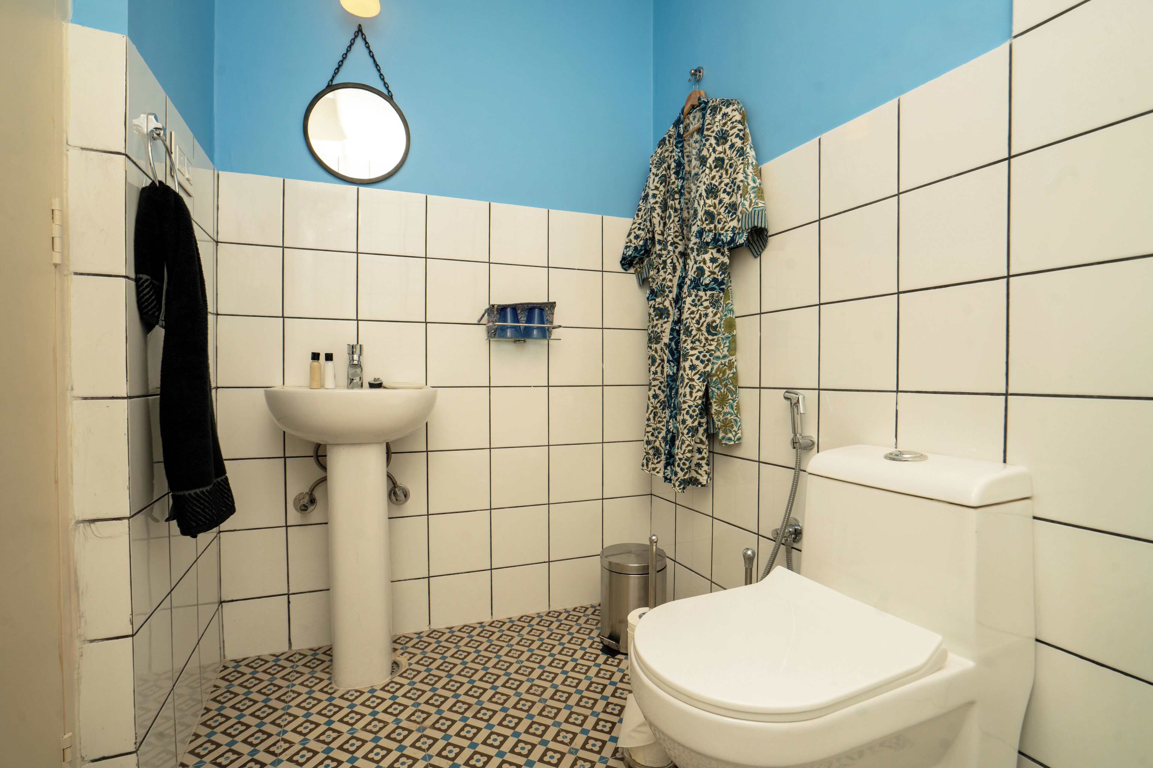 A bathroom with a white sink, toilet, and shower. There is a towel rack with a towel and a hanging robe. A mirror is mounted on the wall above the sink.