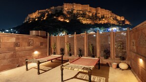 A courtyard with two woven benches, a decorative wall, and a view of a historic fort in the background. - Aakash Heritage Haveli (Jodhpur)