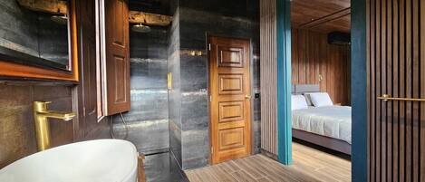 Un baño moderno con una bañera independiente, una puerta de madera y vistas a un dormitorio con cama y mesita de noche.