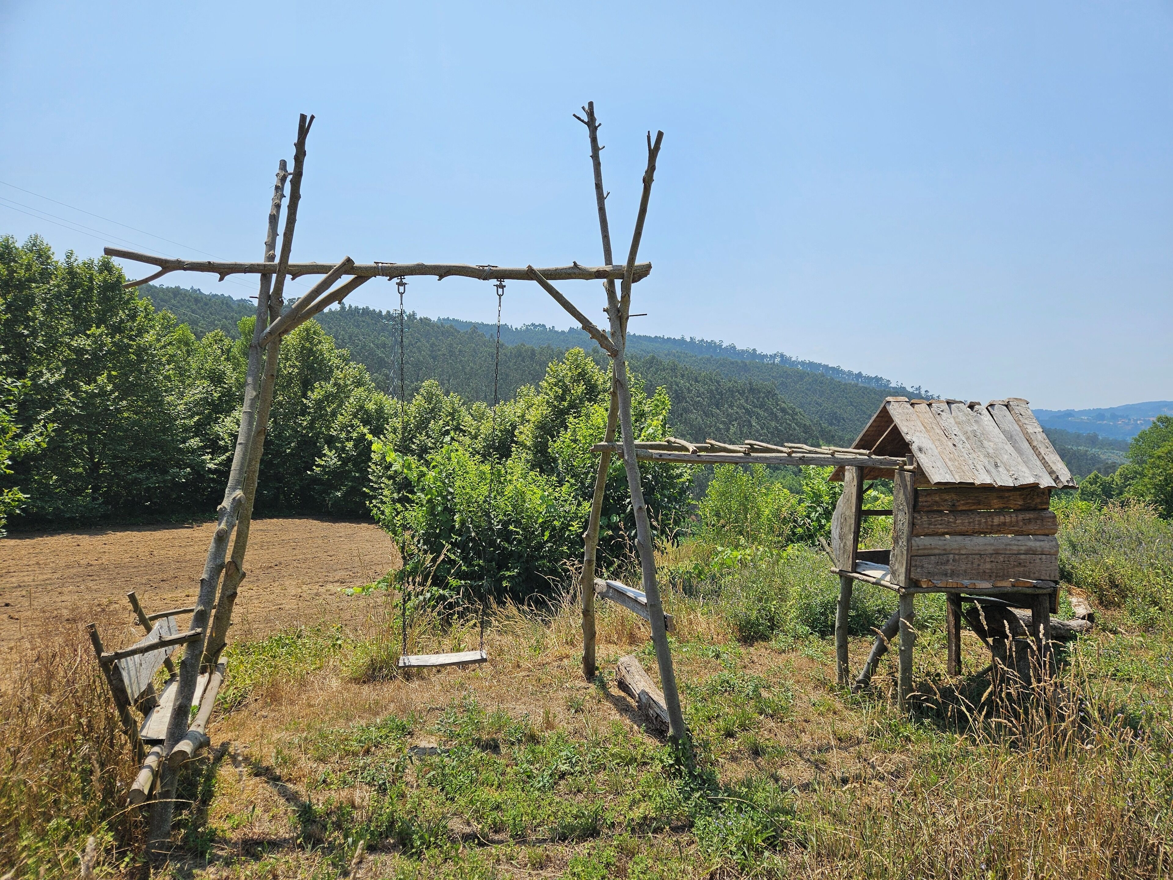 A rustic wooden structure with a slanted roof, surrounded by a natural landscape with trees and hills in the background.