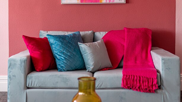 A room with a grey sofa, a colorful abstract painting, and a yellow vase on a white table.