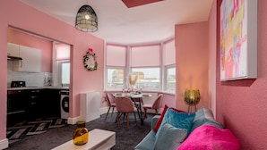 A modern living room with a pink color scheme, a dining area with a table set for two, and a kitchen with a washing machine.
