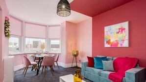 A room with a pink wall, a blue sofa with cushions, a white coffee table, a dining table with chairs, and a window with blinds.