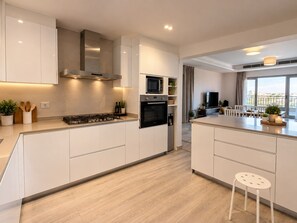 Cocina moderna con muebles blancos, estufa a gas, microondas y horno. Cuenta con un comedor con mesa y sillas, y vistas a la ciudad a través de amplios ventanales.