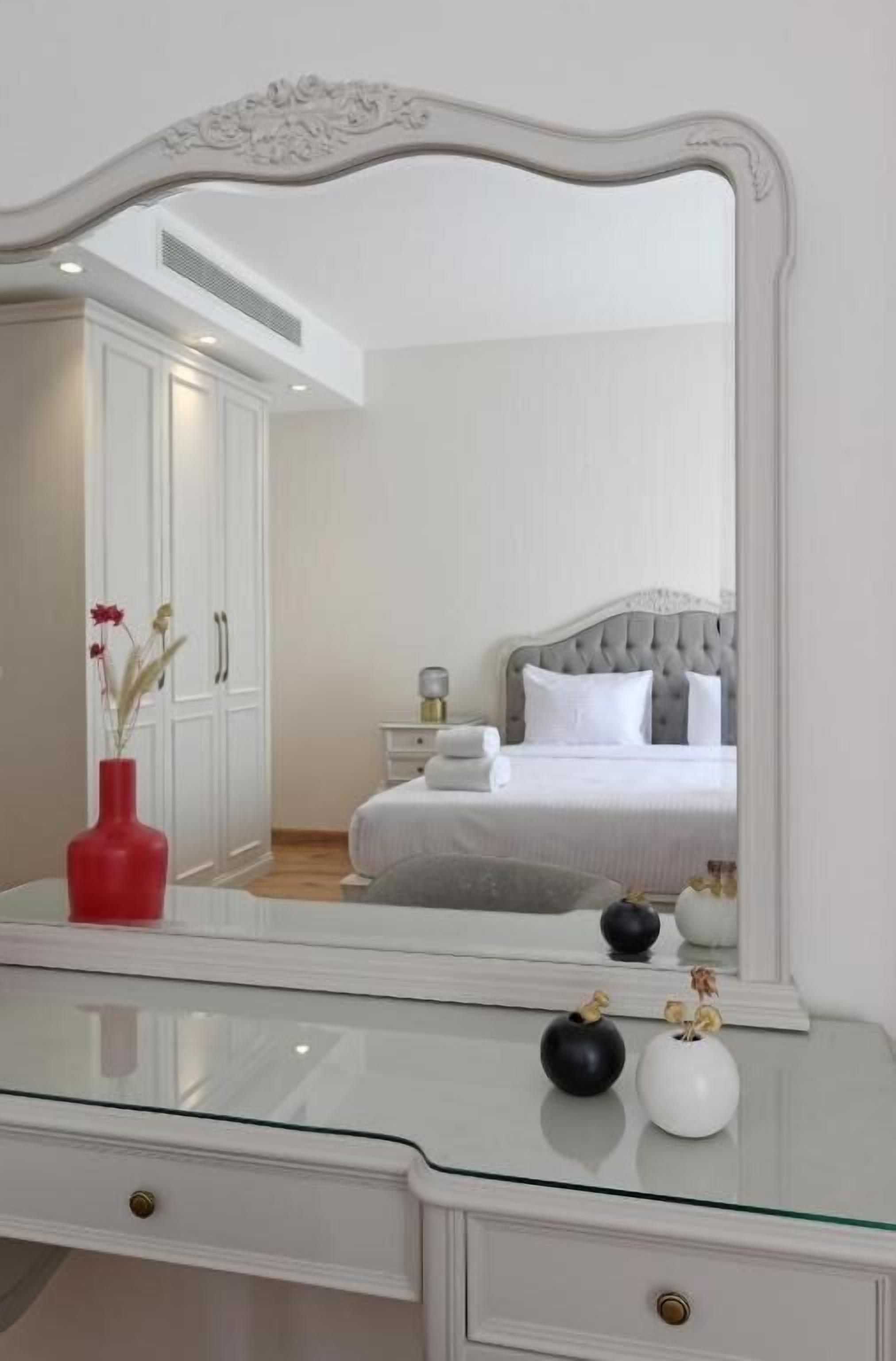 A bedroom with a large mirror, a bed with white bedding, a nightstand, and a dresser with a vase.