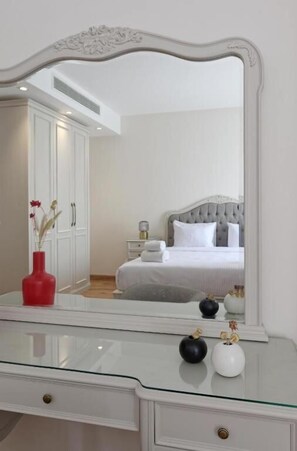 Un dormitorio con un espejo grande, una cama con ropa de cama blanca, una mesita de noche y un aparador con un jarrón.