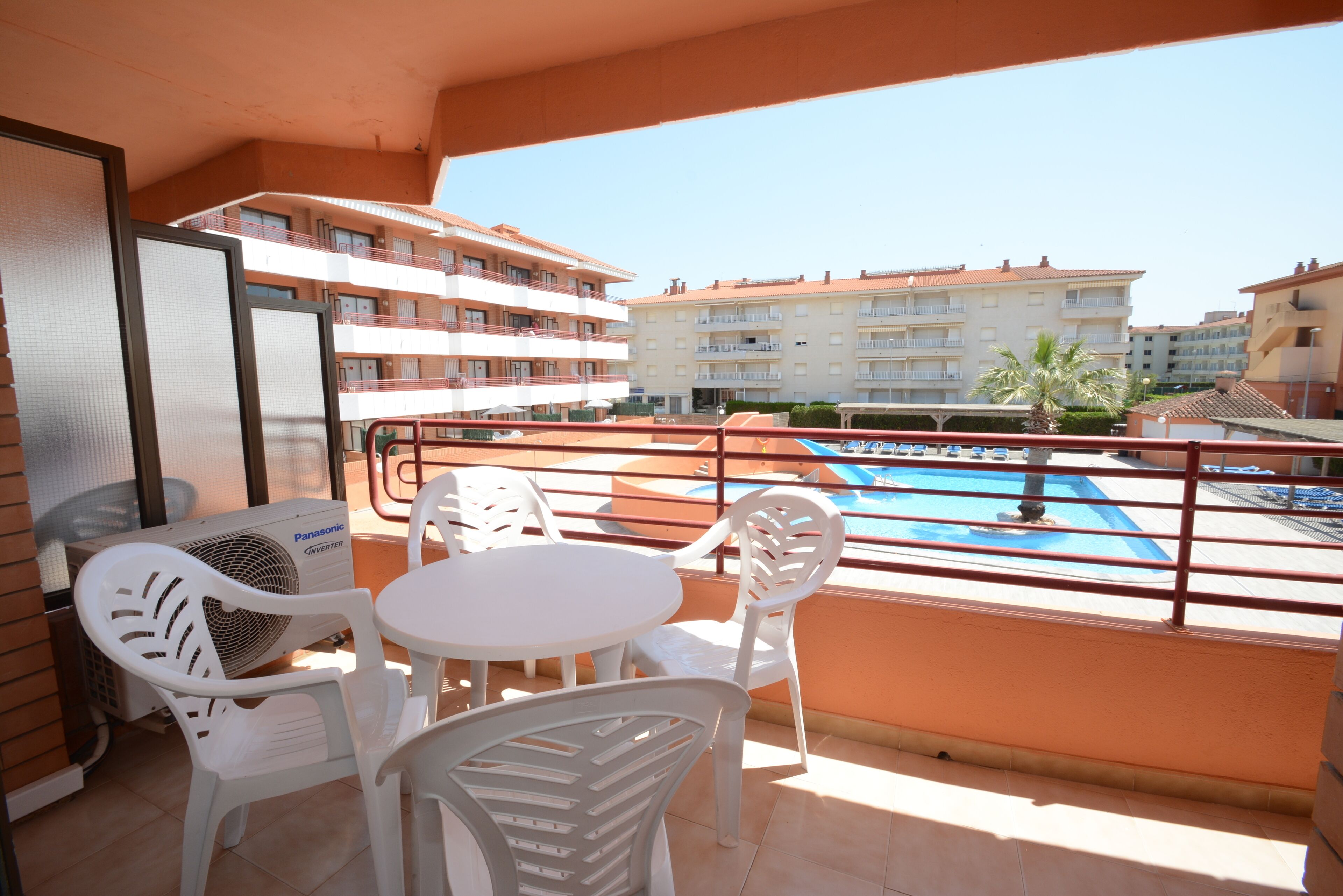 A balcony with a pool view, white plastic chairs, a table, and a Panasonic air conditioner.