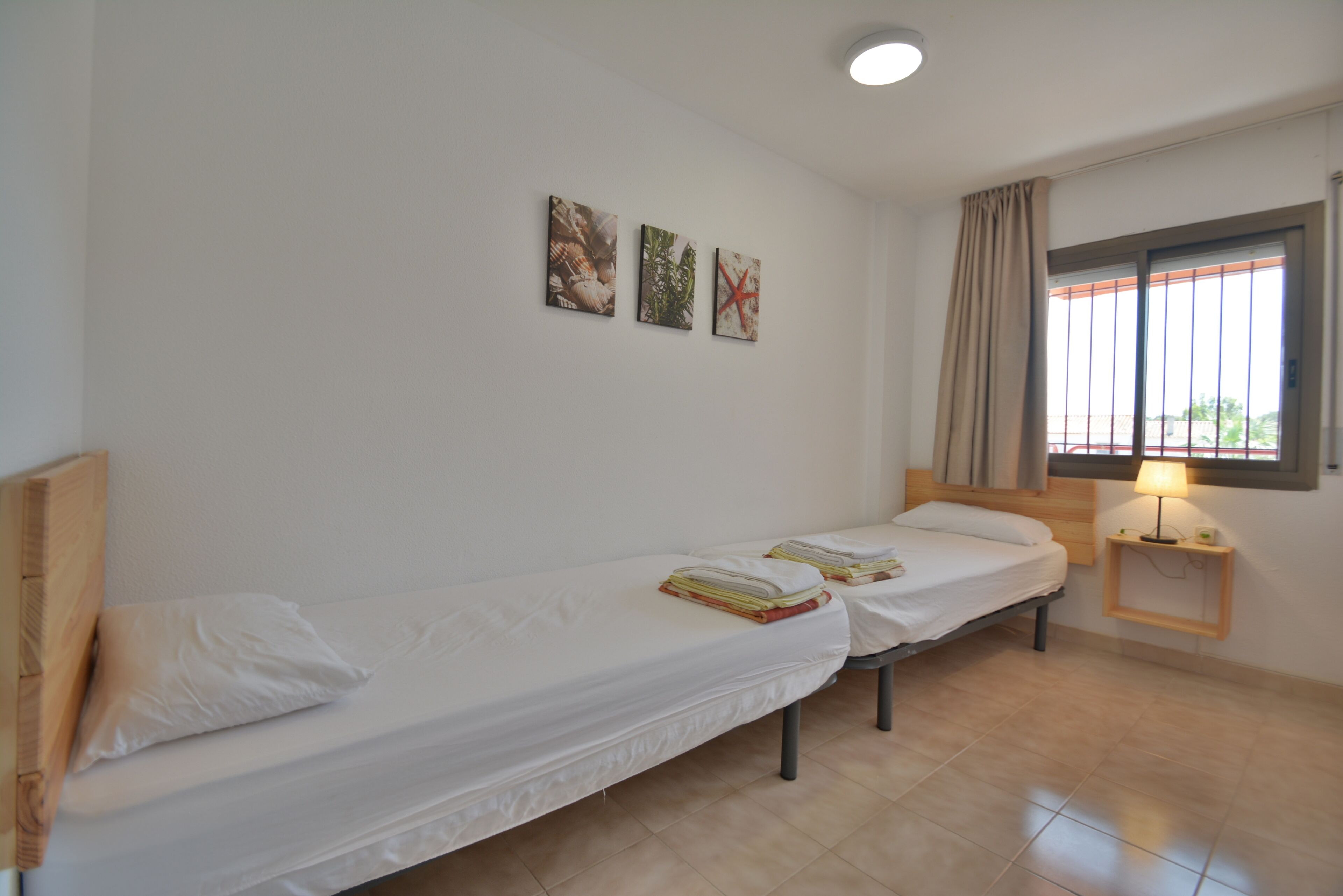 Une chambre avec deux lits simples, une fenêtre avec des rideaux, une table de chevet avec une lampe et trois tableaux accrochés au mur.