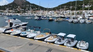 Un port de plaisance où sont amarrés différents types de bateaux, avec une chaîne de montagnes en arrière-plan et des bâtiments le long de la côte.