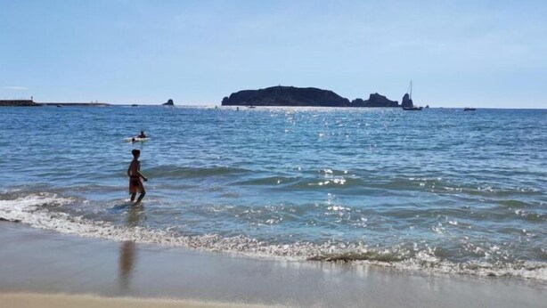 Une plage avec un ciel dégagé, une eau calme et un promontoire rocheux visible au loin.