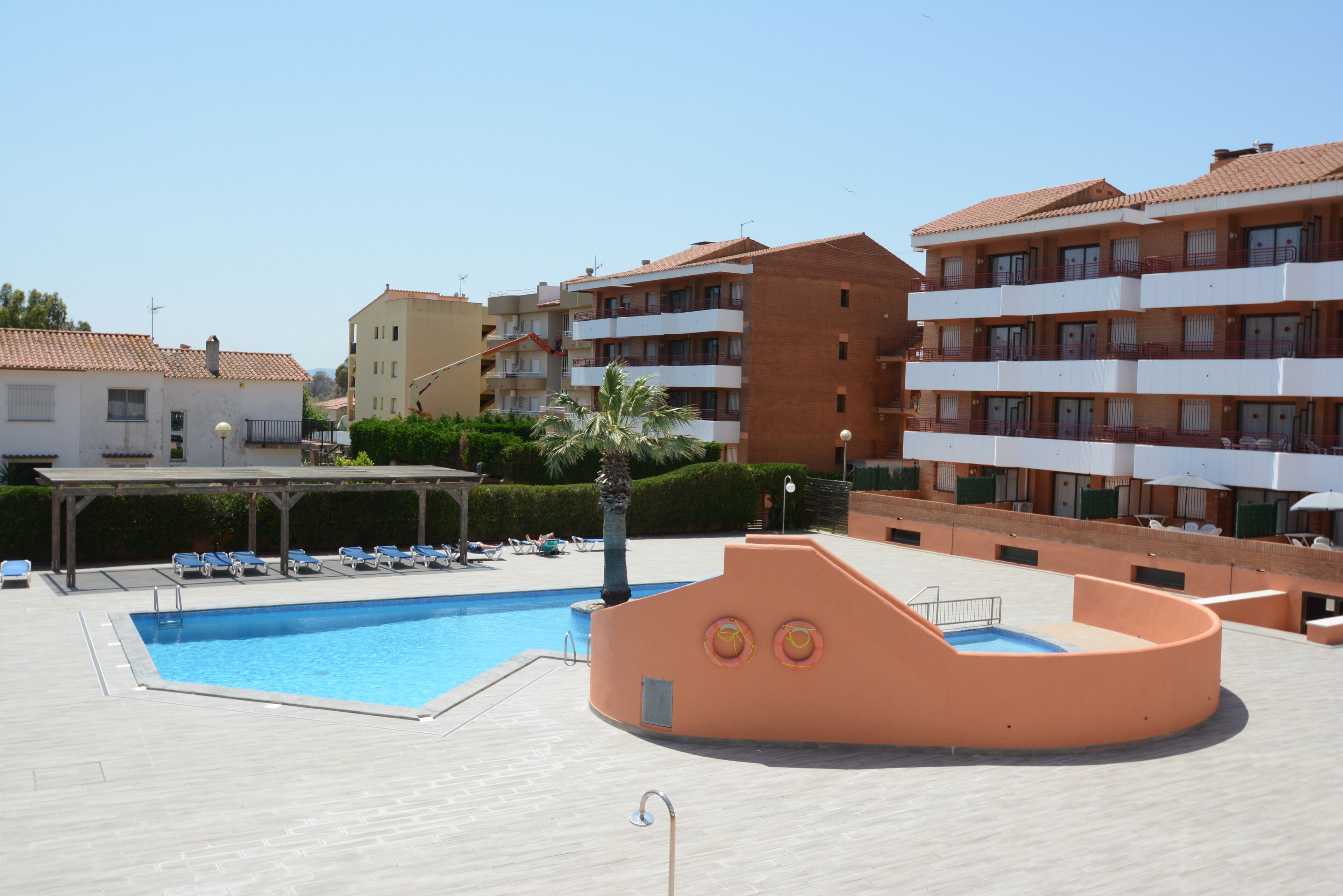 A swimming pool area with lounge chairs, a covered seating area, and a building with multiple balconies.