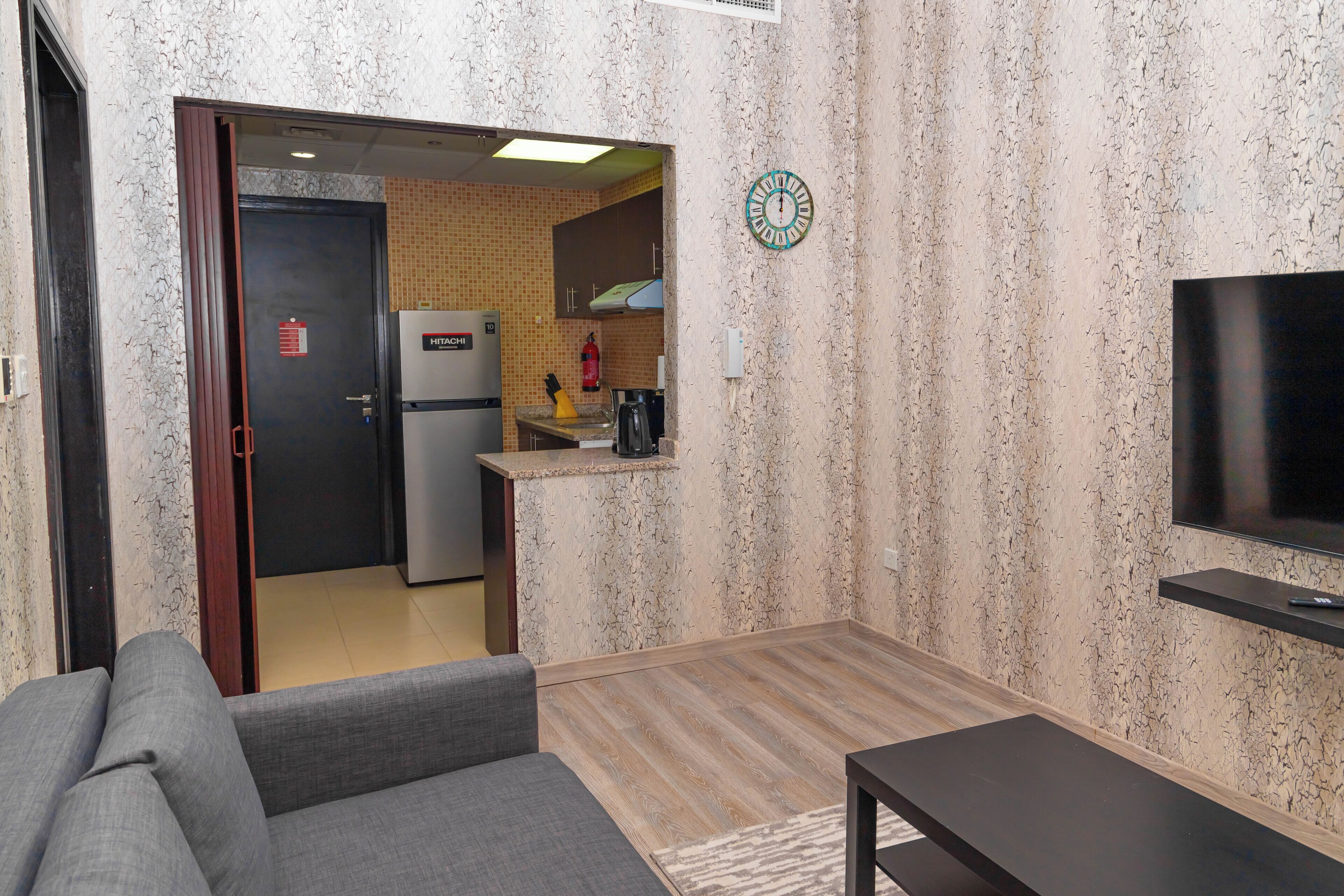A hotel room with a sofa, a TV, a kitchen area, and a clock on the wall.