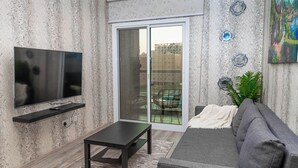 A modern living room with a flat-screen TV, a dark coffee table, a gray sofa, and a sliding glass door leading to a balcony.