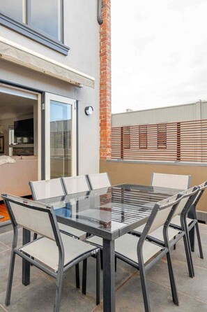 An outdoor patio with a glass-top table and six chairs, a brick column, and a view of a building with a metal fence.