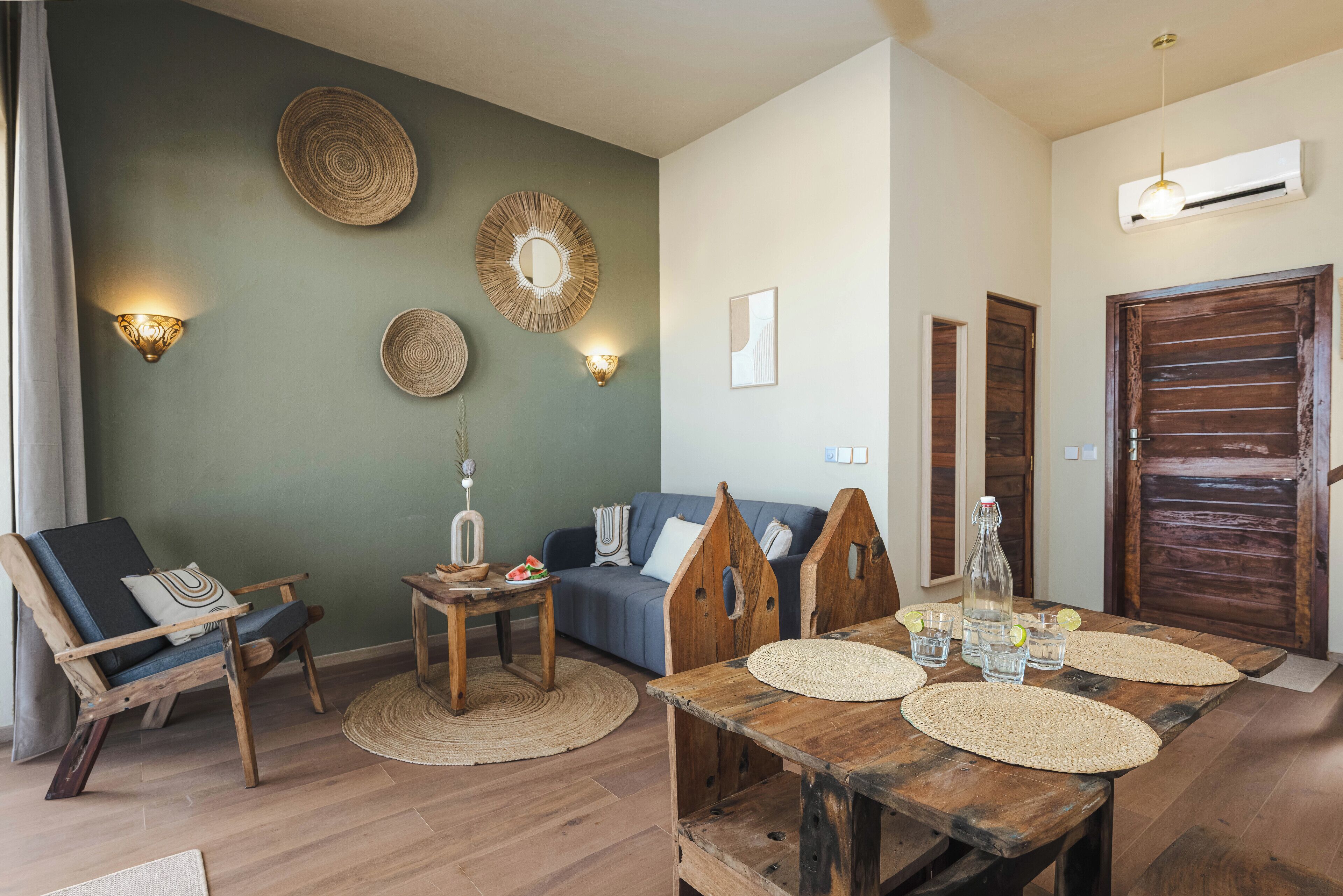 A cozy living room with a wooden dining table, a blue sofa, and a wooden chair. There are wall-mounted lights and a large mirror.