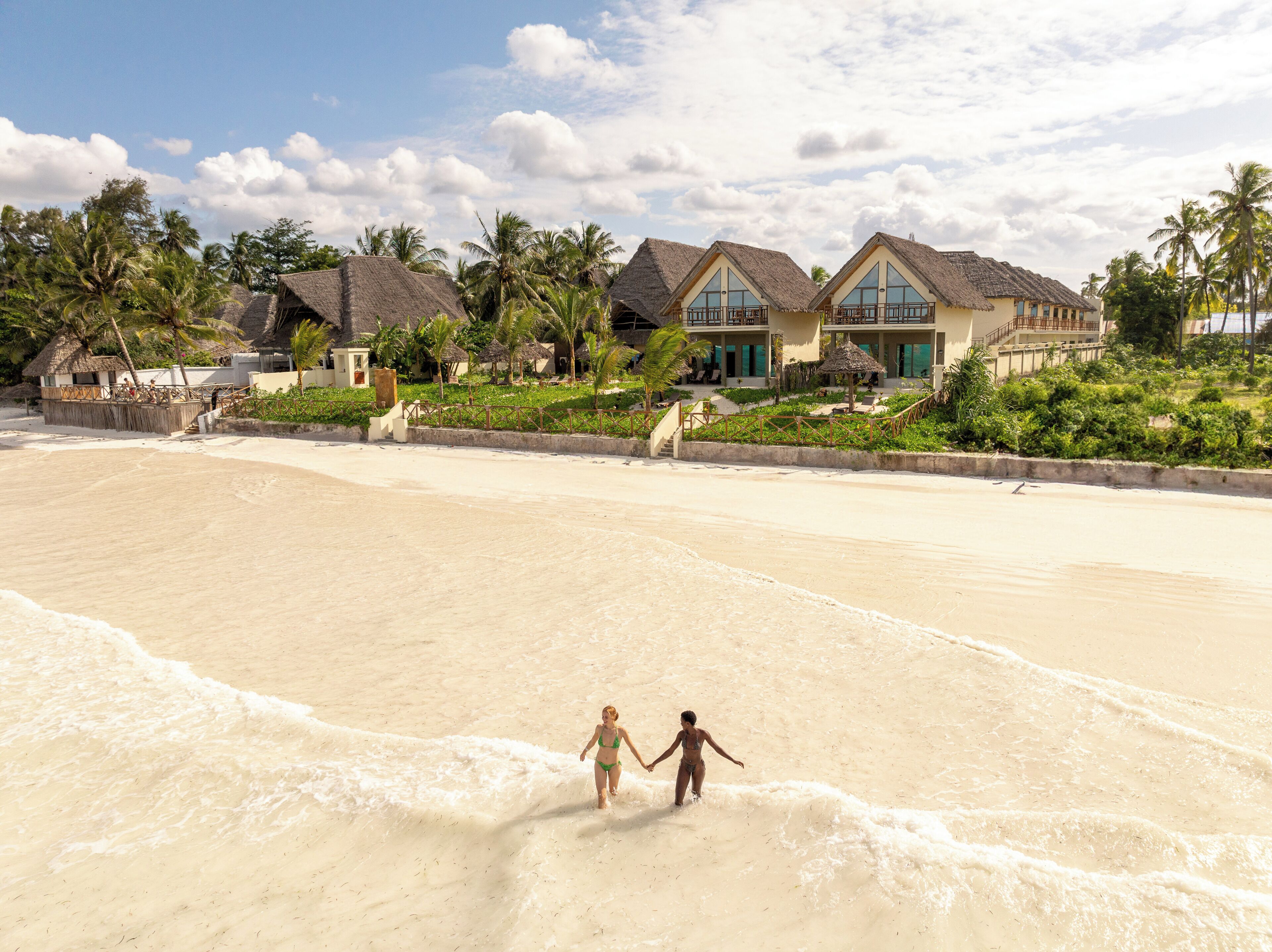 Un resort frente al mar con villas de techo de paja, una playa arenosa y palmeras.