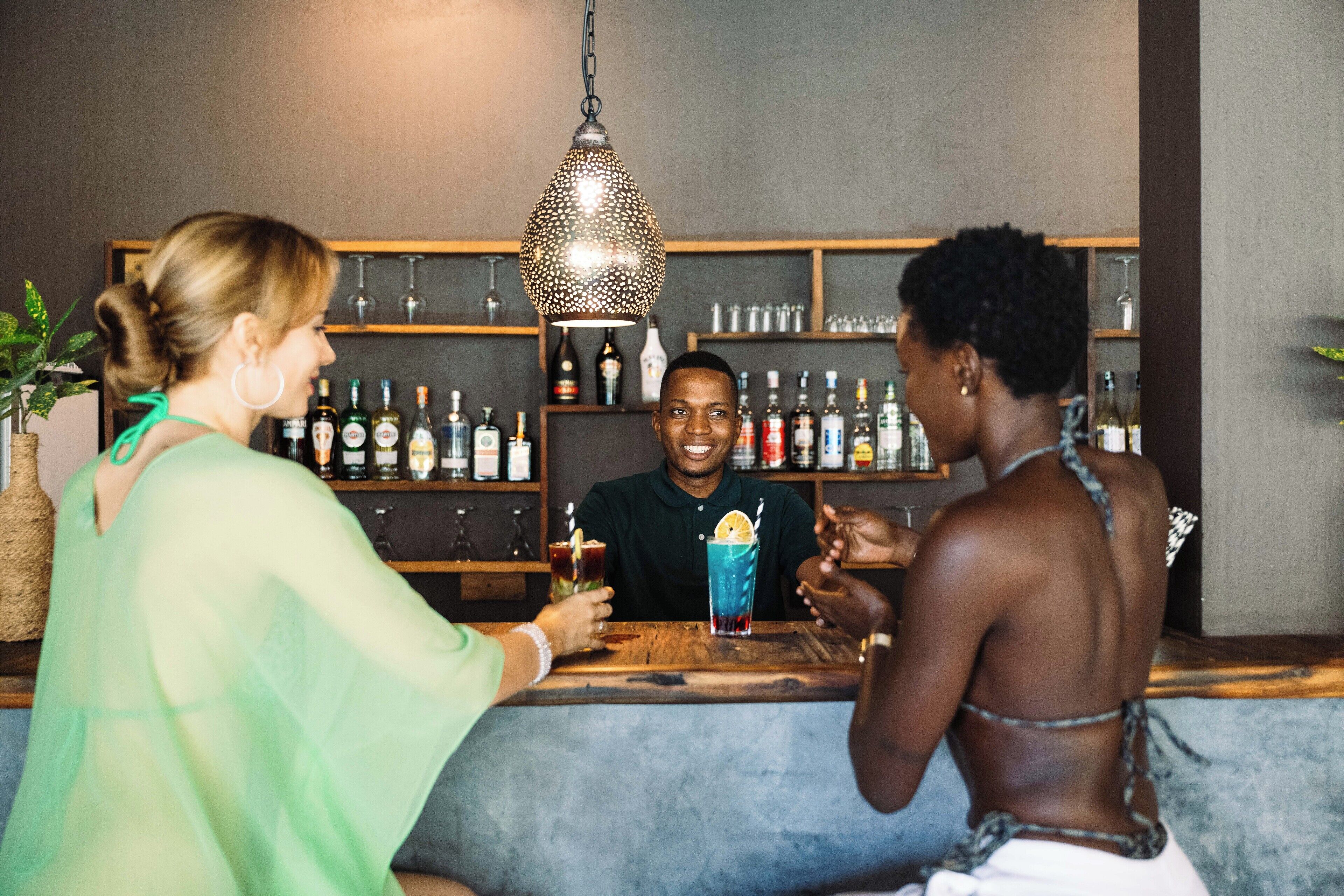 Una zona de bar con mostrador de madera, estantes con botellas y una lámpara colgante.